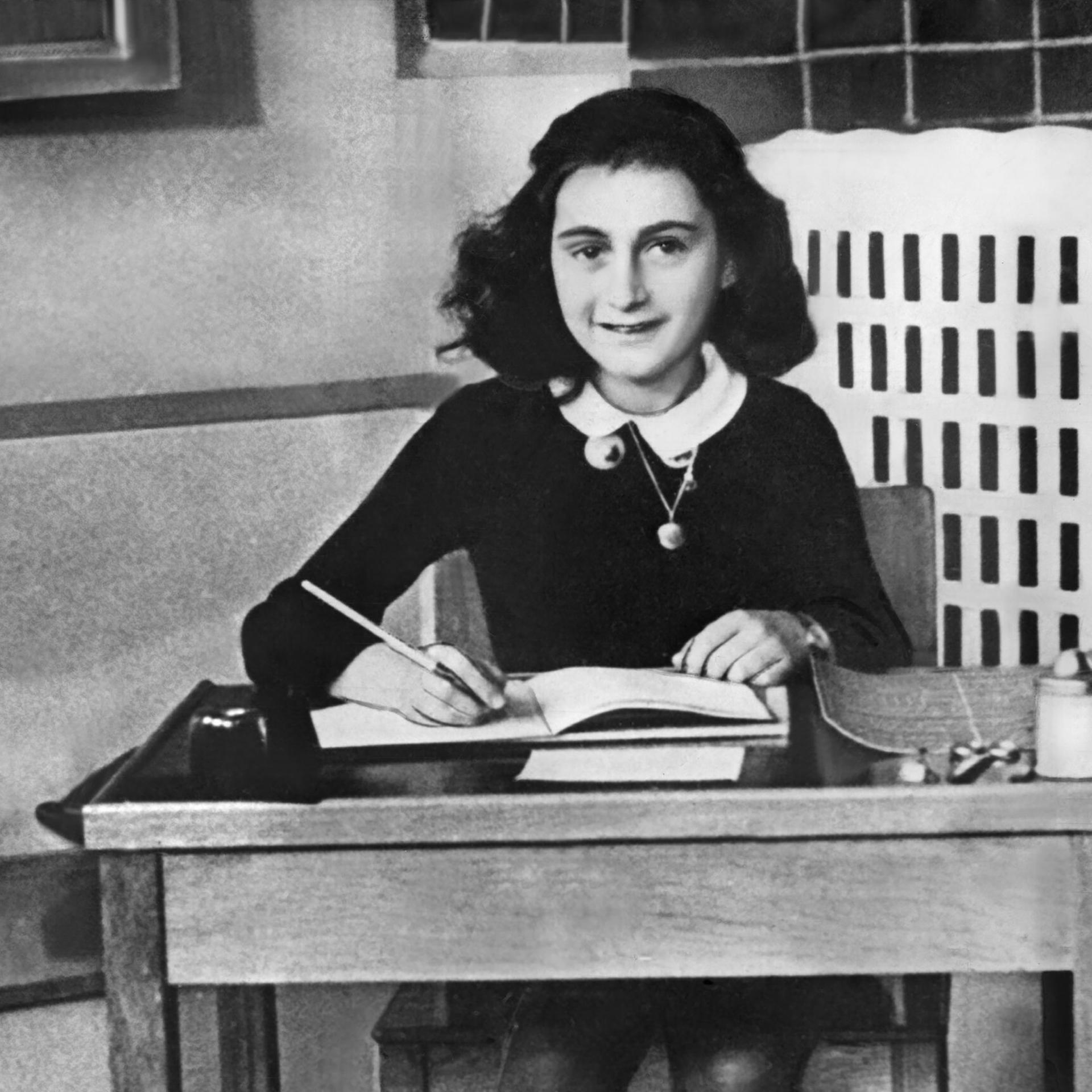 Schwarz-Weiß-Fotografie von Anne Frank, die an einem hölzernen Schreibtisch sitzt und lächelnd von ihrem Tagebuch aufschaut, in das sie schreibt.