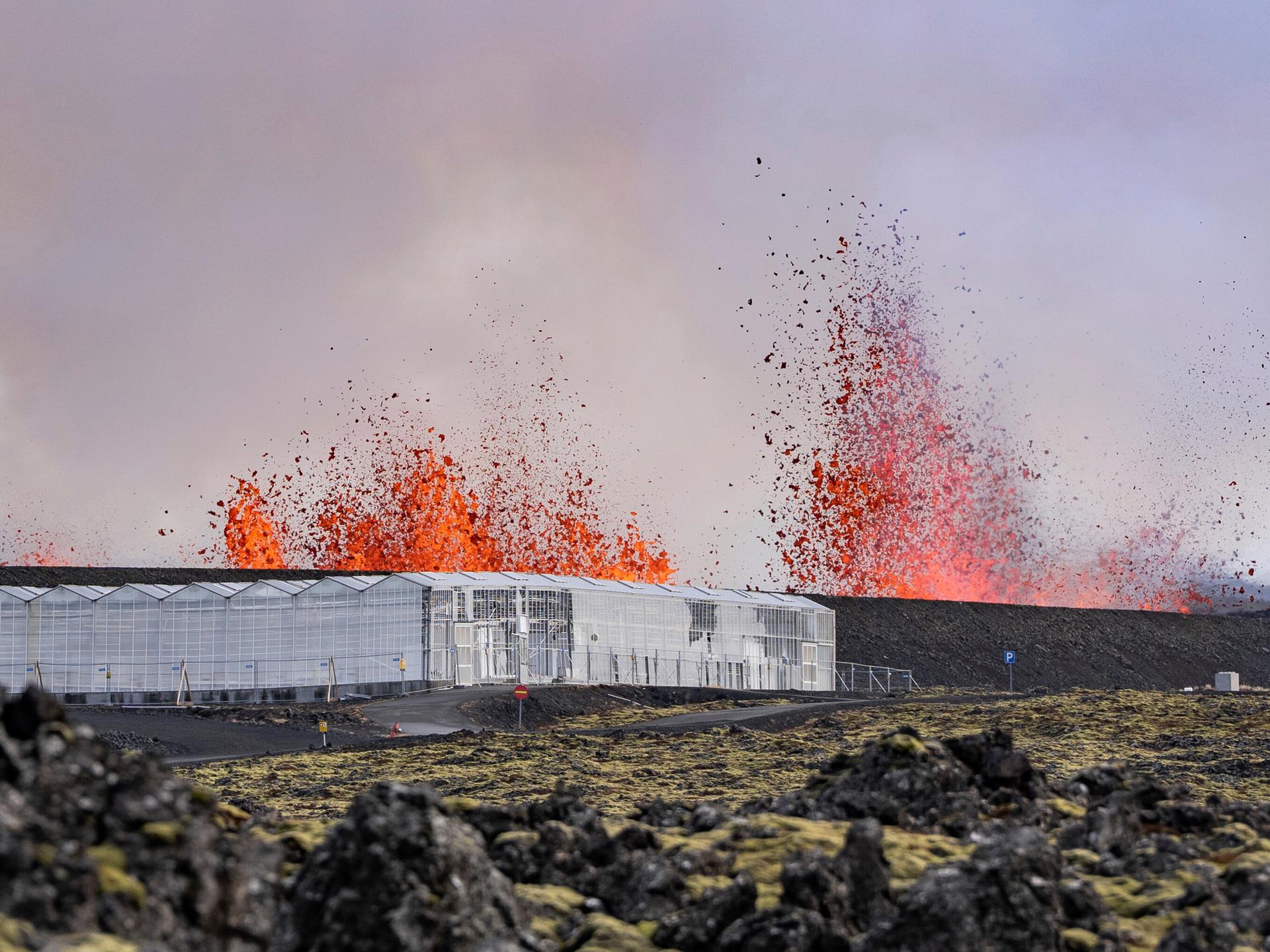 Vulkanausbruch in der Nähe von Grindavik. Im Vordergrund ein Gebäude aus Glas, das auf bergartig zerklüftetem Untergrund steht. Im Hintergrund lodern tiefrote Flammen, die von einem Vulkan stammt. 