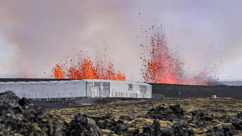 Vulkanausbruch in der Nähe von Grindavik. Im Vordergrund ein Gebäude aus Glas, das auf bergartig zerklüftetem Untergrund steht. Im Hintergrund lodern tiefrote Flammen, die von einem Vulkan stammt. 