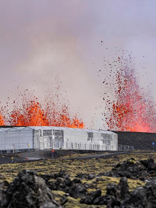 Vulkanausbruch in der Nähe von Grindavik. Im Vordergrund ein Gebäude aus Glas, das auf bergartig zerklüftetem Untergrund steht. Im Hintergrund lodern tiefrote Flammen, die von einem Vulkan stammt. 