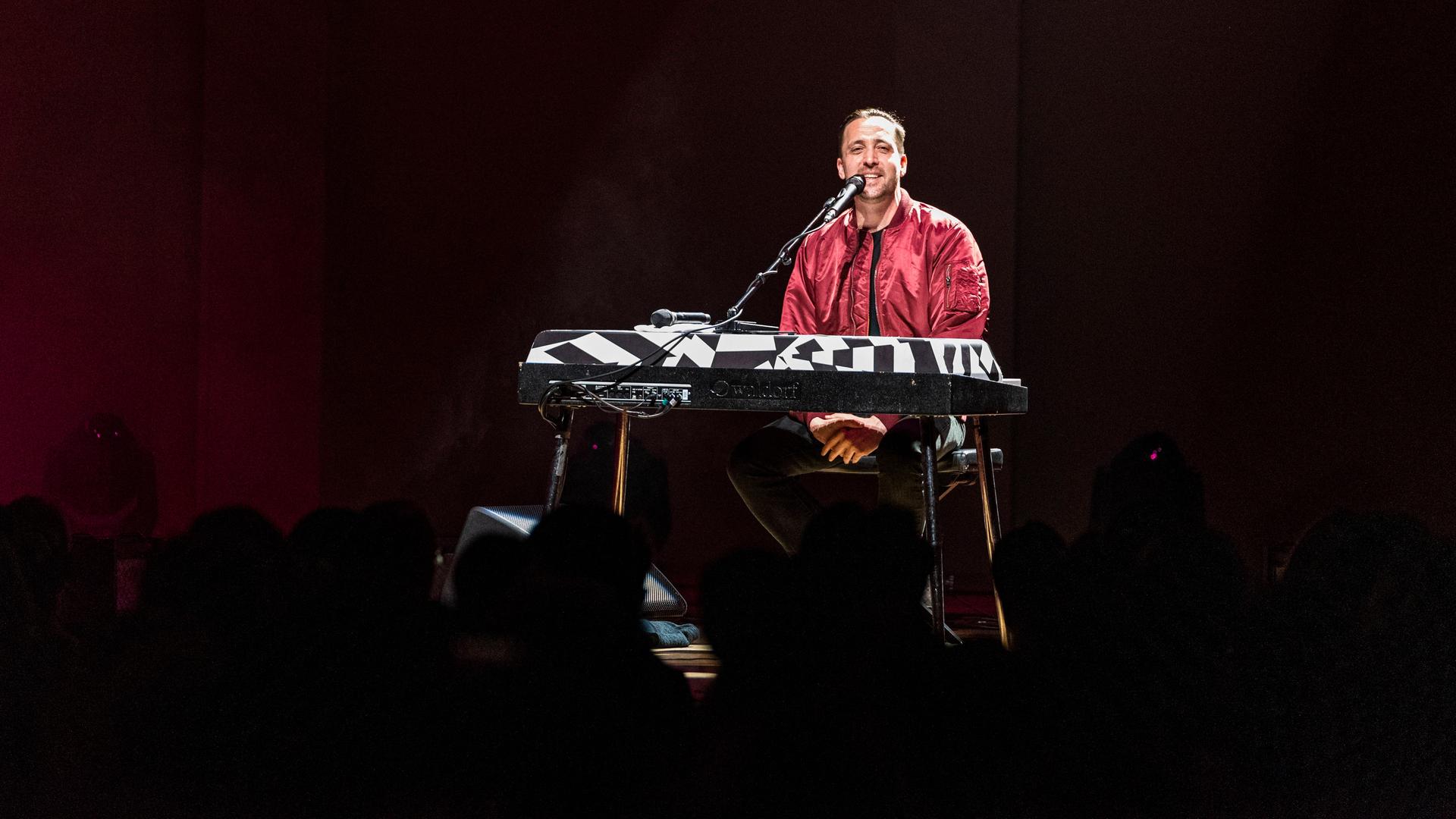 Danger Dan sitzt auf einer dunklen Bühne vor einem Keyboard und einem Mikrofon. Danger Dan sitzt auf einer dunklen Bühne vor einem Keyboard und einem Mikrofon.