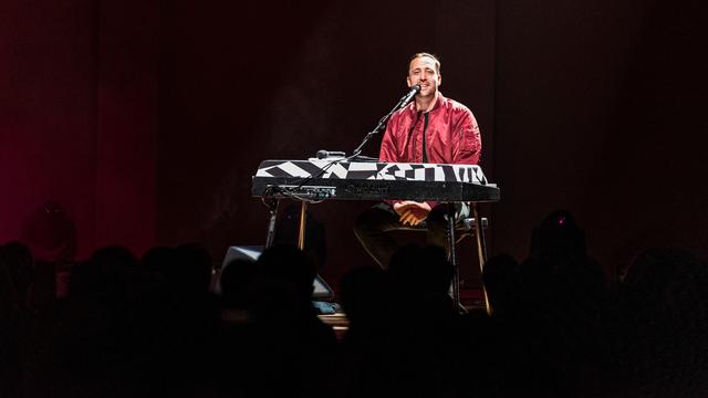 Danger Dan sitzt auf einer dunklen Bühne vor einem Keyboard und einem Mikrofon.