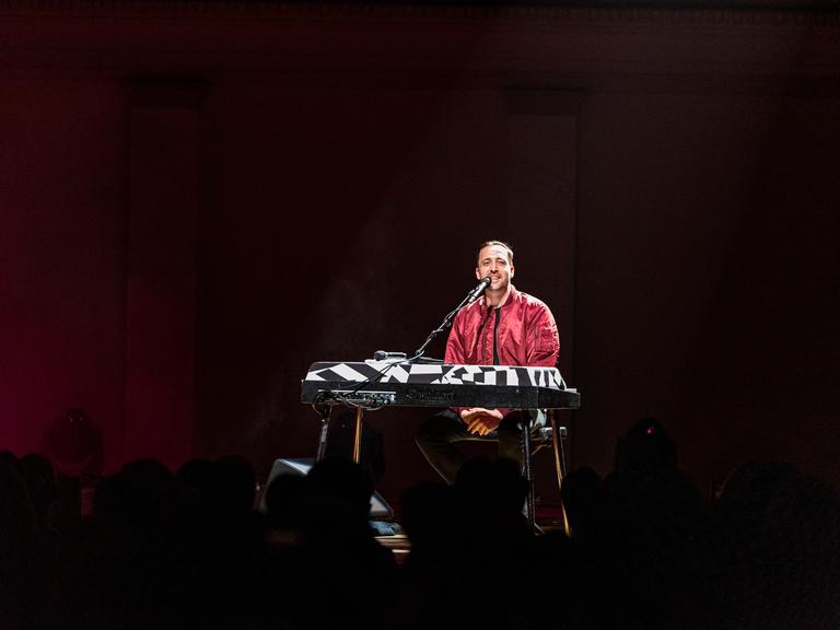 Danger Dan sitzt auf einer dunklen Bühne vor einem Keyboard und einem Mikrofon.