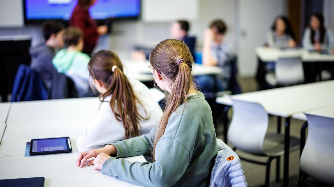 Schülerinnen und Schüler einer siebten Klasse hören in einem Klassenraum ihrer Lehrerin zu. Sie sitzen an verschiedenen Schultischen.