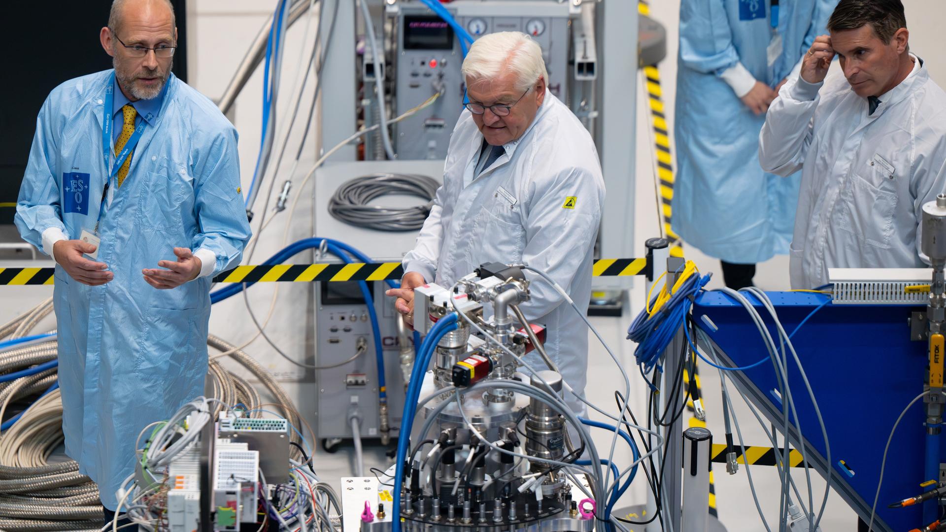 Bundespräsident Frank-Walter Steinmeier besucht die Technikhalle der Europäischen Südsternwarte. Steinmeier trägt einen weißen Kittel und steht vor einem Apparat, aus dem Kabel herausführen. 