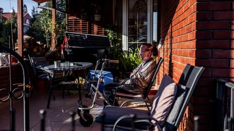 Eine ältere Frau sitzt auf der Terrasse eines Wohnhauses. Vor ihr steht ein Rollator.