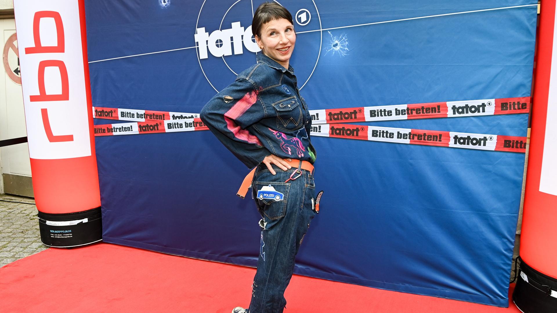 Schauspielerin Meret Becker steht ganz in Jeans gekleidet mit bunten Applikationen vor einer blauen Stellwand mit dem Tatort-Logo und rot-weißem Absperrband mit der Aufschrift "Tatort - Bitte betreten!". An den Seiten sind je eine Säule aufgestellt, auf denen das Logo RBB prankt. Schauspielerin Meret Becker steht ganz in Jeans gekleidet mit bunten Applikationen vor einer blauen Stellwand mit dem Tatort-Logo und rot-weißem Absperrband mit der Aufschrift "Tatort - Bitte betreten!". An den Seiten sind je eine Säule aufgestellt, auf denen das Logo RBB prankt.
