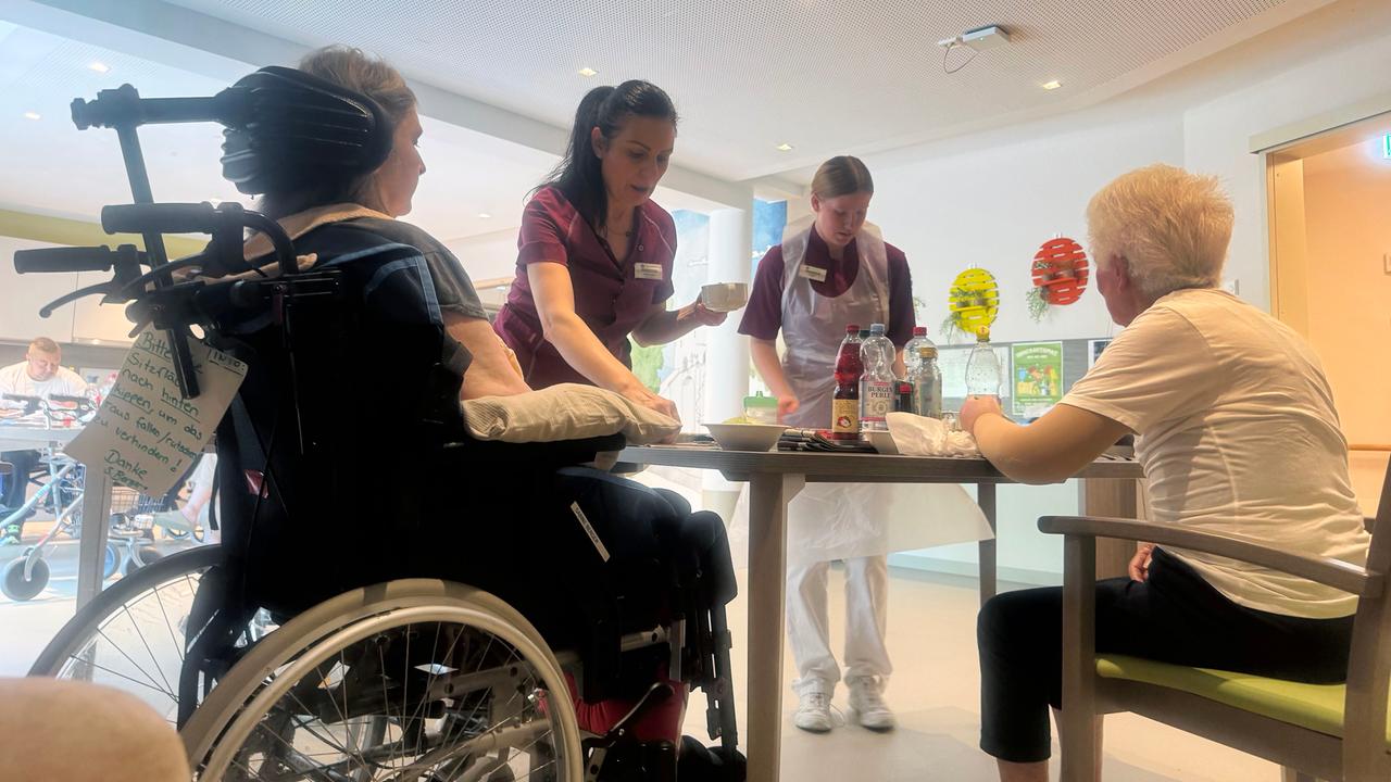 Senioren sitzen an einem Tisch und werden von Pflegern mit Essen versorgt.
