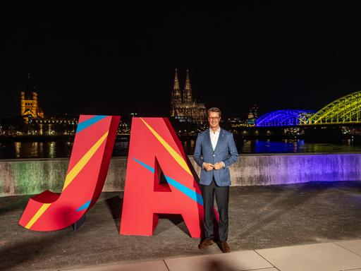 NRW-Ministerpräsident Hendrik Wüst steht nachts am Rheinufer vor großen roten Buchstaben „JA“, mit dem beleuchteten Kölner Dom und einer in Olympiafarben angestrahlten Rheinbrücke im Hintergrund