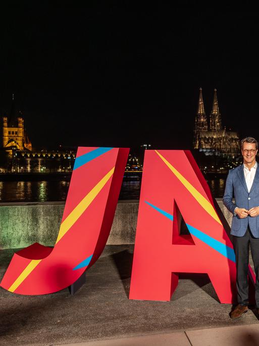 NRW-Ministerpräsident Hendrik Wüst steht nachts am Rheinufer vor großen roten Buchstaben „JA“, mit dem beleuchteten Kölner Dom und einer in Olympiafarben angestrahlten Rheinbrücke im Hintergrund