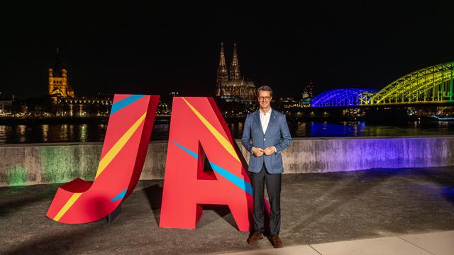 NRW-Ministerpräsident Hendrik Wüst steht nachts am Rheinufer vor großen roten Buchstaben „JA“, mit dem beleuchteten Kölner Dom und einer in Olympiafarben angestrahlten Rheinbrücke im Hintergrund