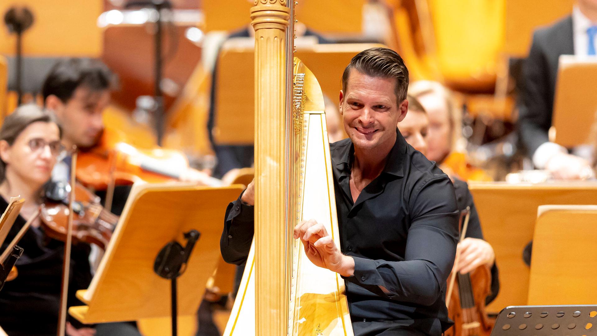 Ein Mann sitzt in schwarzer Konzertkleidung an einer goldenen Harfe. Die Hände an den Saiten. Er lächelt. Hinter ihm sind einzelne Orchestermusiker zu erkennen. 