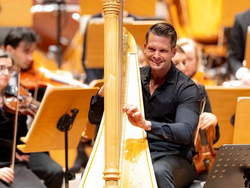 Ein Mann sitzt in schwarzer Konzertkleidung an einer goldenen Harfe. Die Hände an den Saiten. Er lächelt. Hinter ihm sind einzelne Orchestermusiker zu erkennen. 
