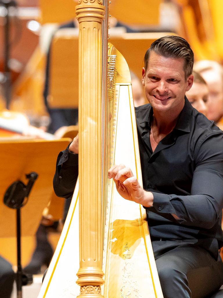 Ein Mann sitzt in schwarzer Konzertkleidung an einer goldenen Harfe. Die Hände an den Saiten. Er lächelt. Hinter ihm sind einzelne Orchestermusiker zu erkennen. 