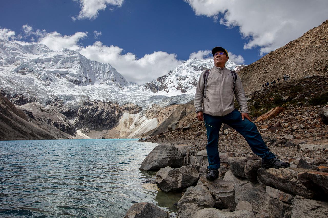 Saúl Luciano Lliuya steht auf Felsen neben dem Gletschersee Palcacocha, im Hintergrund sind eis- und schneebedeckte Berge Saúl Luciano Lliuya steht auf Felsen neben dem Gletschersee Palcacocha, im Hintergrund sind eis- und schneebedeckte Berge