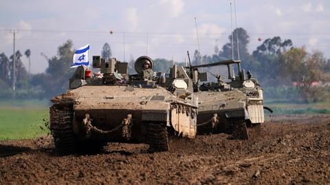 Man sieht einen Panzer und dahinter ein anderes gepanzertes Fahrzeug durch eine schlammige Fläche fahren. Im Hintergrund unscharf eine Baumreihe.