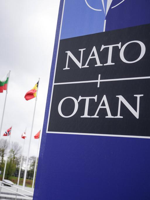 Fahnen der NATO-Mitgliedsstaaten vor dem Brüsseler NATO-Hauptquartier neben einem Schild mit der Aufschrift NATO.