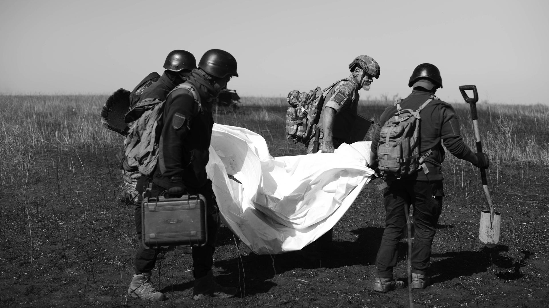 Vier Personen mit Schutzausrüstung tragen die Überreste eines gefallenen Soldaten unter einer weißen Plane über ein Feld auf diesem schwarz-weiß Foto.