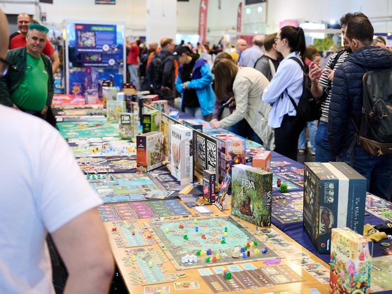 Besucher stehen beim Medien-Rundgang der internationalen Publikumsmesse "Spiel Essen 2025“ in der Messe Essen um einen langen Tisch, auf dem verschiedene Spiele präsentiert werden. 