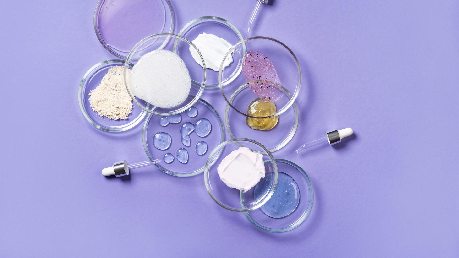 Auf einem violetten Untergrund stehen mehrere Petrischalen in denen verschiedene Kosmetika enthalten sind.