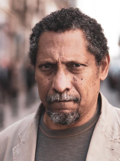 Percival Everett steht auf eines Straße und schaut in die Kamera. Er hat kurze, angegraute, lockige Haare und trägt einen Kinnbart. Zu einem braun Shirt trägt er ein helles Sakko.