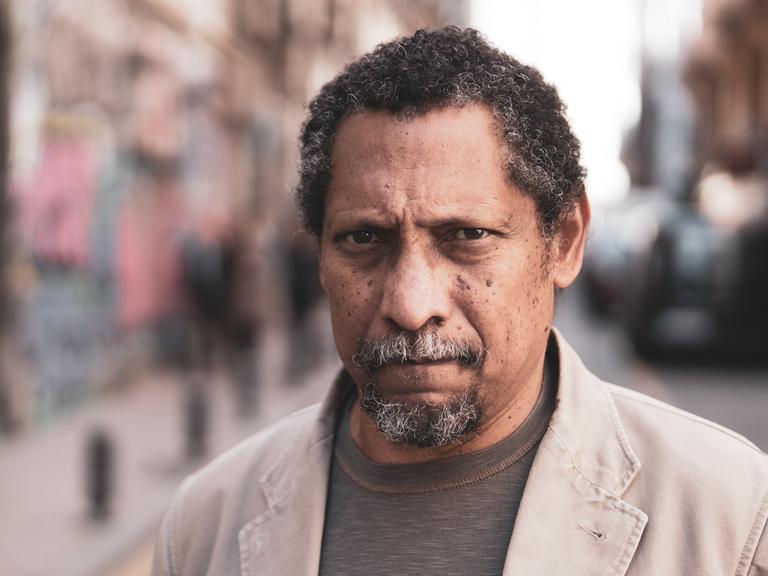 Percival Everett steht auf eines Straße und schaut in die Kamera. Er hat kurze, angegraute, lockige Haare und trägt einen Kinnbart. Zu einem braun Shirt trägt er ein helles Sakko.