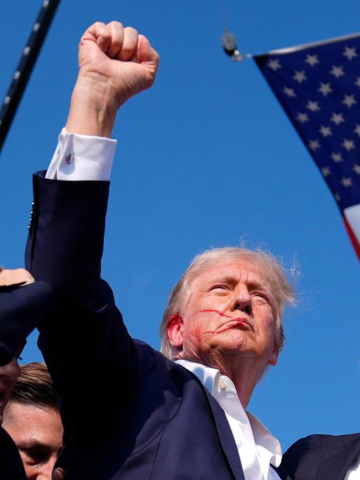 Ex-US-Präsident Donald Trump reckt die Faust in den Himmel. An seinem rechten Ohr ist Blut zu sehen. Im Hintergrund: Die US-Flagge