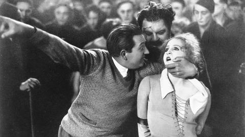 Ein Schwarz-Weiß-Foto zeigt, wie Fritz Lang (links) am Set von "Metropolis" Brigitte Helm (rechts) festhält. Zwischen den beiden ist Heinrich George zu sehen. 