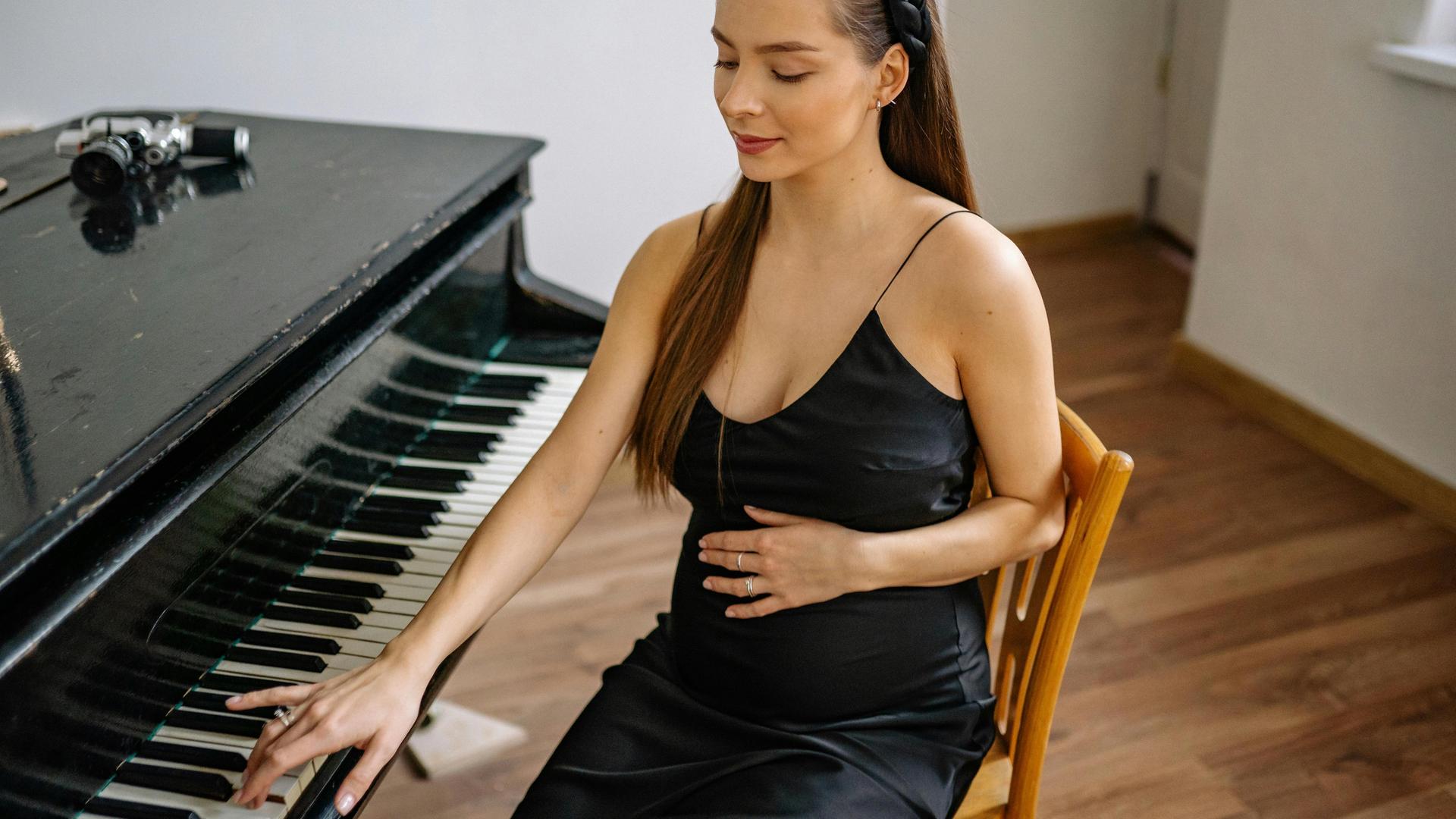 Eine schwangere Frau sitzt neben einem Flügel, eine Hand ist sanft auf ihren Bauch gelegt, die andere berührt die Tasten des Instrumentes.