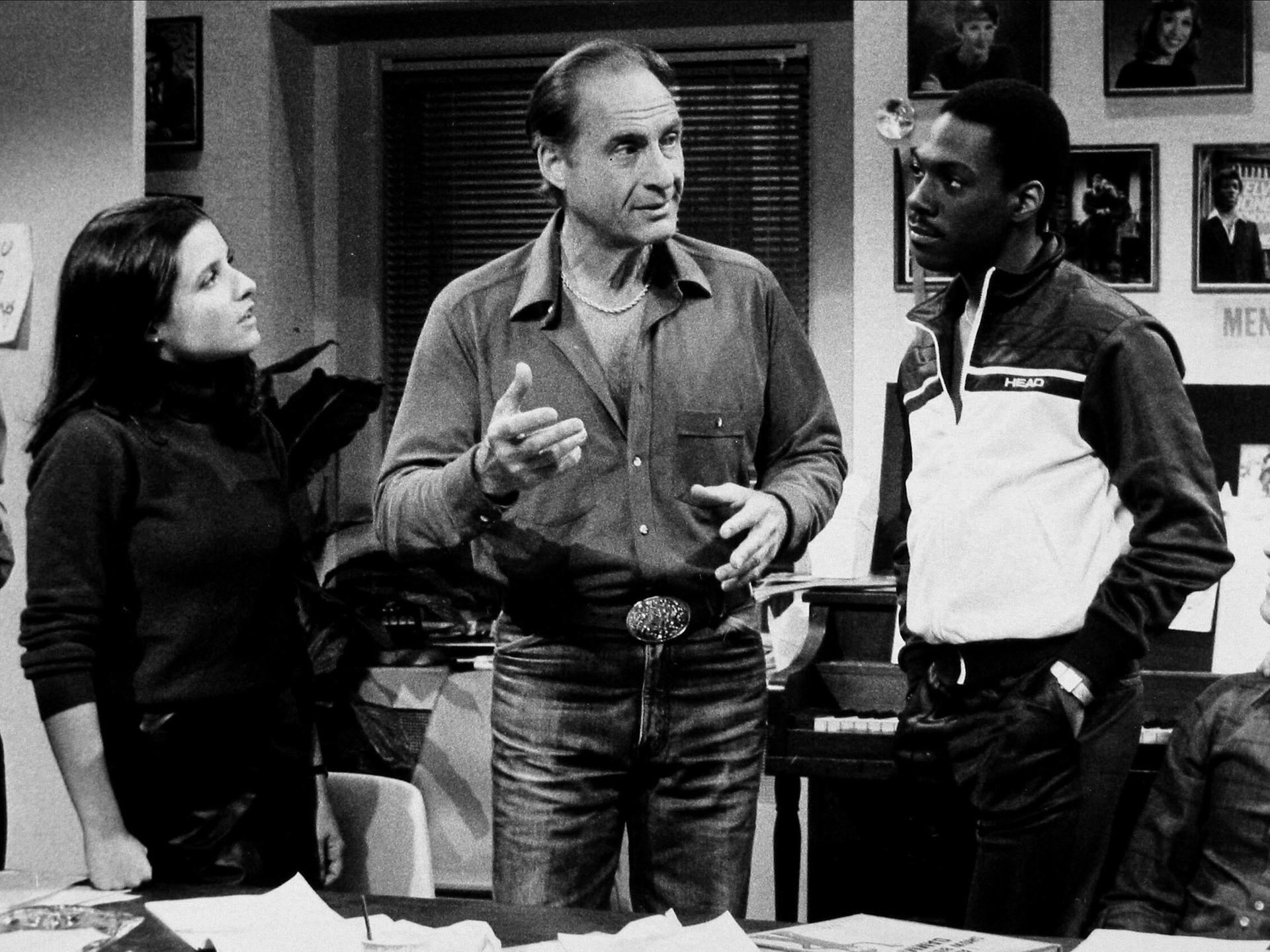 Ein Schwarz-Weiß-Foto zeigt die US-Komiker Julia Louis Dreyfus, Sid Caesar und Eddie Murphy (v. l.n.r.) in einer Ausgabe von „Saturday Night Live“ im Jahr 1983.
