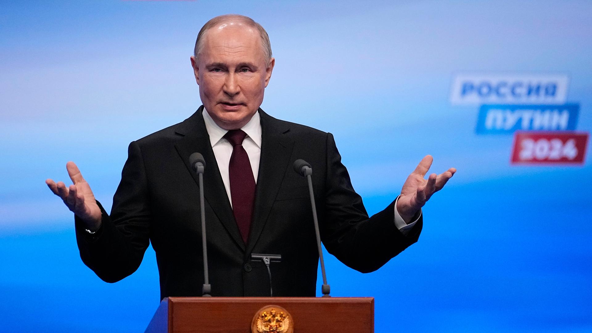 Wladimir Putin spricht an einem Rednerpult in zwei Mikrofone und hebt die Arme.