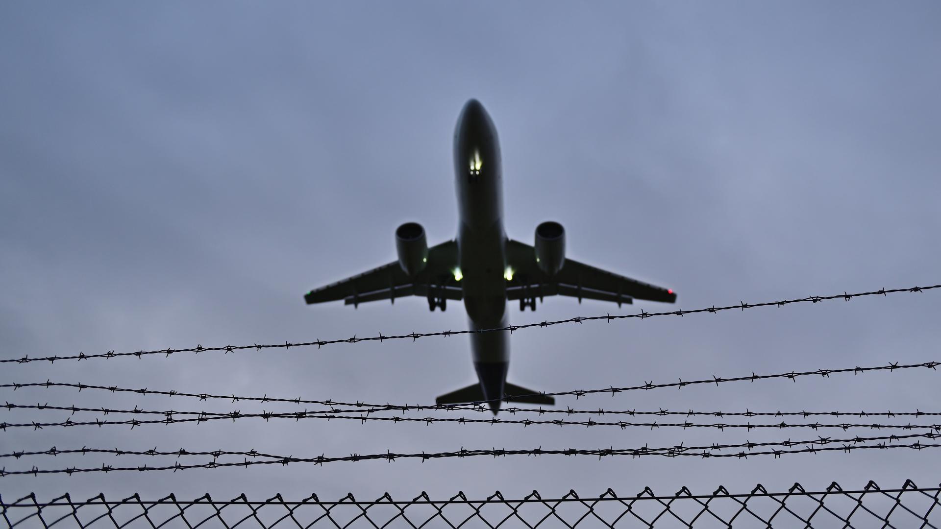 Ein Flugzeug fliegt über einen Stacheldraht-Zaun. Der Himmel ist grau.