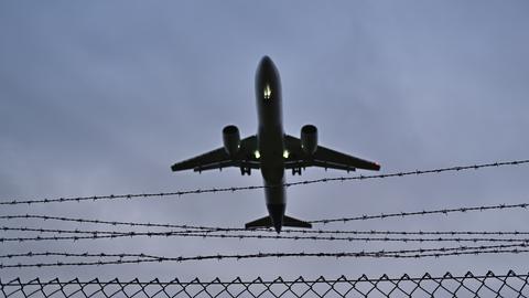 Blick in den grauen Himmel: Ein Flugzeug fliegt über einem Stacheldrahtzaun.