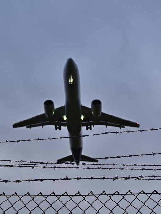 Blick in den grauen Himmel: ein Flugzeug fliegt über einem Stacheldrahtzaun