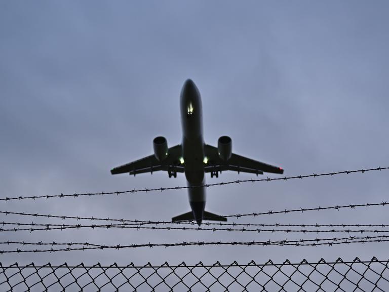 Blick in den grauen Himmel: ein Flugzeug fliegt über einem Stacheldrahtzaun