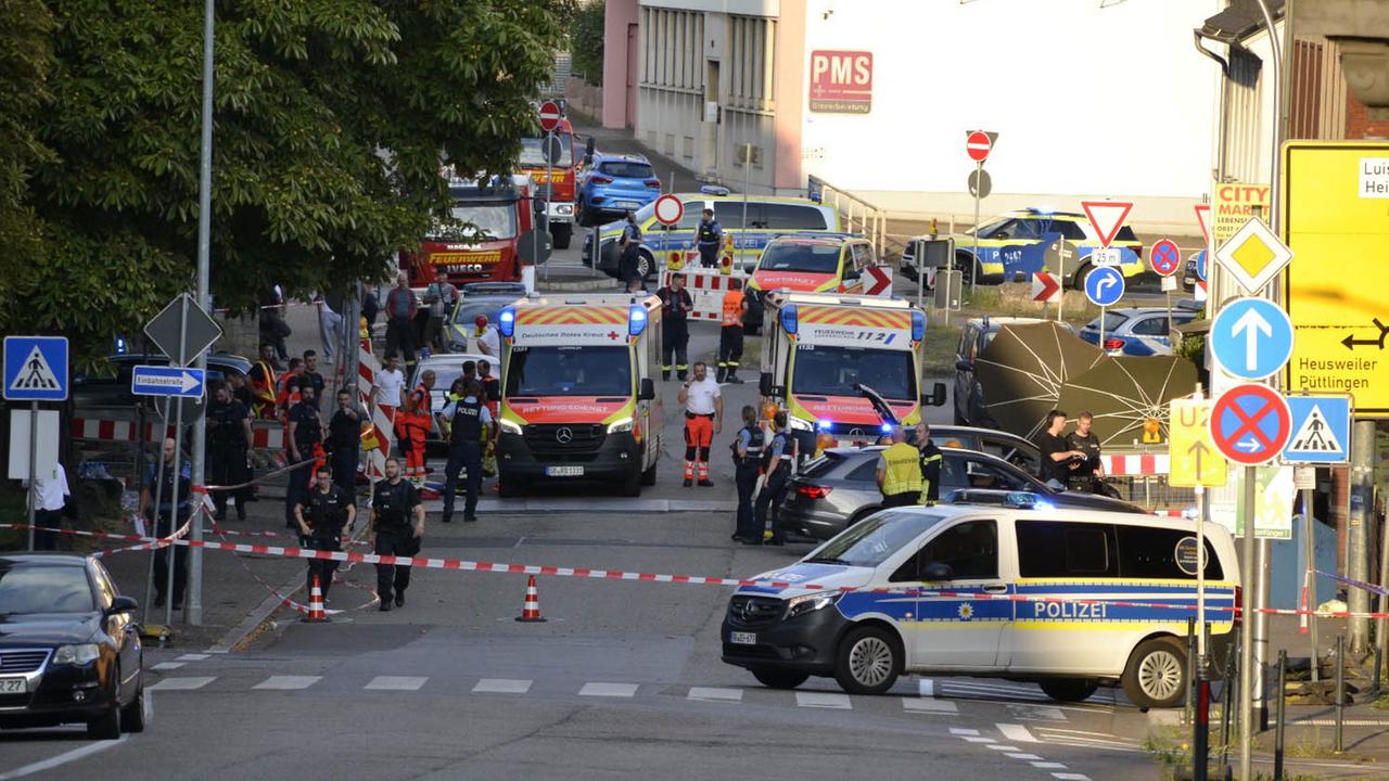 Blick aus der Ferne auf eine mit Flatterband abgesperrte Straße. Auf ihr stehen ein Polizeifahrzeug und zwei Rettungswagen. Polizisten und Sanitäter gehen herum.
