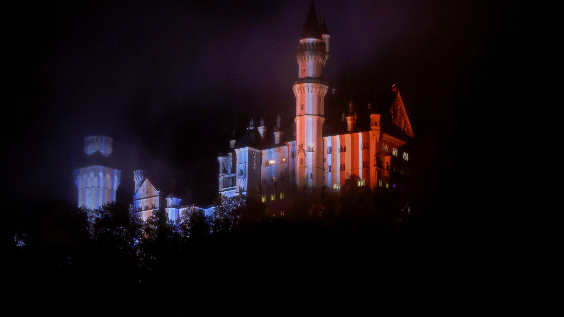 Schwangau: Blick auf das in Blau- und Rottöne beleuchtete Schloss Neuschwanstein. Das Schloss wird in den Farben der bayerischen Klimastreifen beleuchtet.
