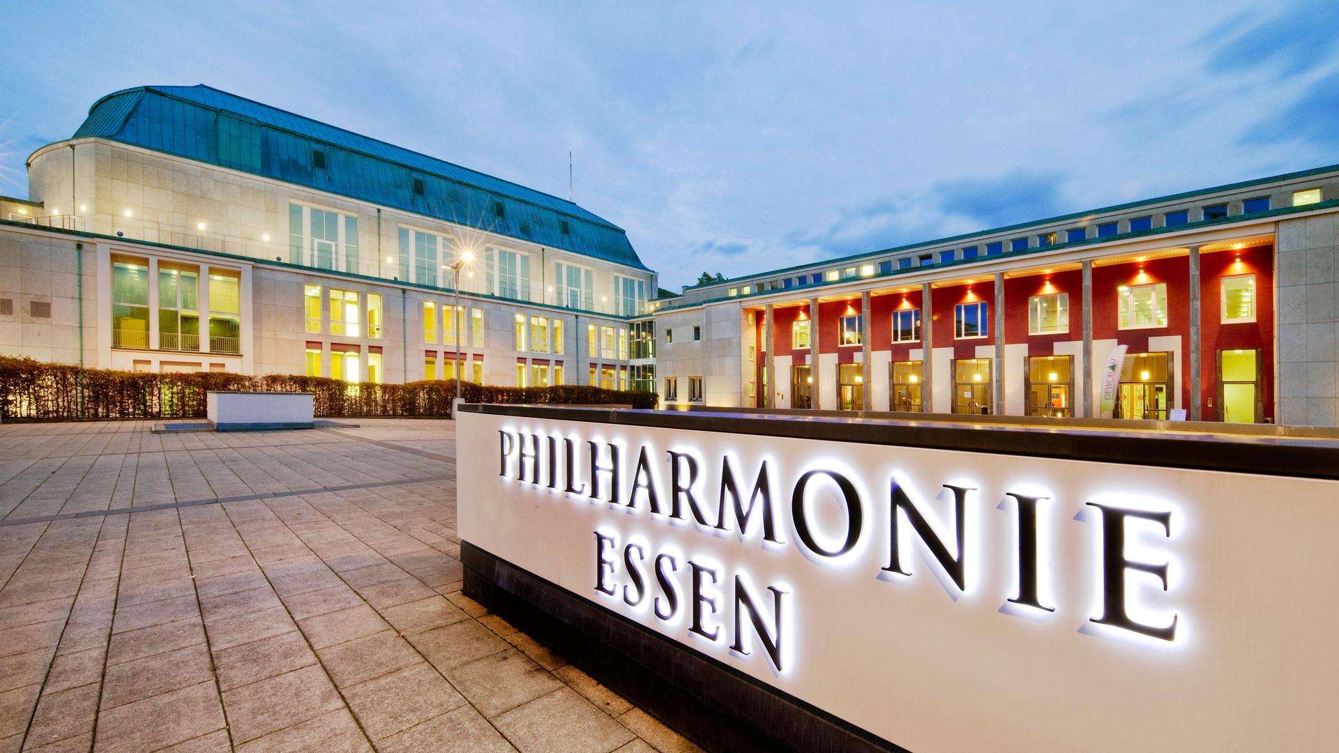 Im Vordergrund leuchtet der Schriftzug der Philharmonie Essen. Im Hintergrund steht das erleuchtete Konzerthaus. Im Vordergrund leuchtet der Schriftzug der Philharmonie Essen. Im Hintergrund steht das erleuchtete Konzerthaus.