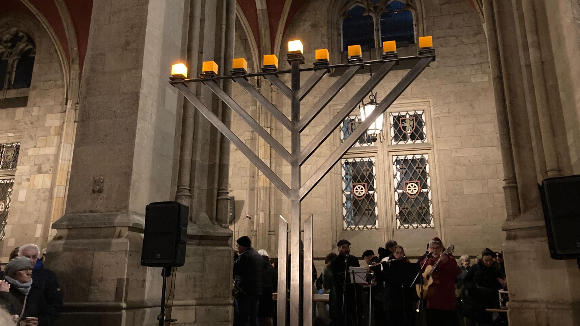 Menschen verfolgen das Entzünden von dem 1. Lichts am Chanukka-Leuchter vor dem Erfurter Rathaus. Sie machen Musik. Auf dem Leuchter brennen zwei Lichter. Menschen verfolgen das Entzünden von dem 1. Lichts am Chanukka-Leuchter vor dem Erfurter Rathaus. Sie machen Musik. Auf dem Leuchter brennen zwei Lichter.