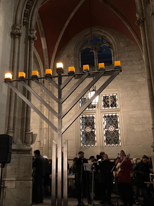 Menschen verfolgen das traditionelle Entzünden des ersten Lichts am Chanukka-Leuchter vor dem Erfurter Rathaus. Sie machen Musik. Auf dem Leuchter brennen zwei Lichter.