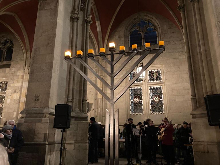 Menschen verfolgen das traditionelle Entzünden des ersten Lichts am Chanukka-Leuchter vor dem Erfurter Rathaus. Sie machen Musik. Auf dem Leuchter brennen zwei Lichter.