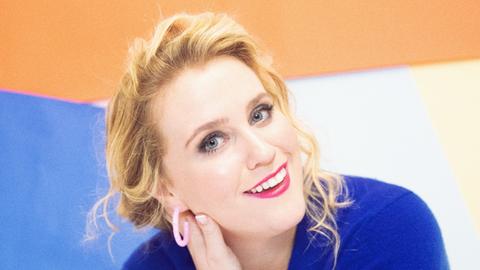 Eine Frau mit blonden Locken posiert lächelnd vor buntem Hintergrund.