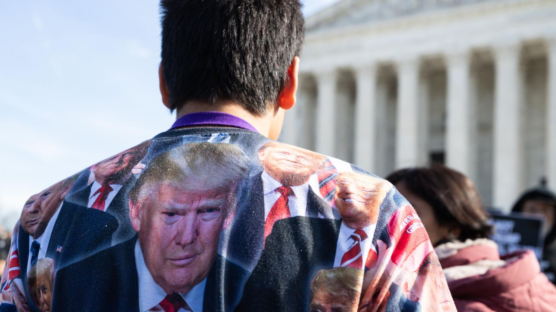 Ein Trump-Anhänger steht vor dem Supreme-Court. Er trägt ein Shirt voller Gesicher von Donald Trump.