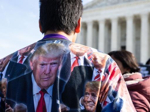 Ein Trump-Anhänger steht vor dem Supreme-Court. Er trägt ein Shirt voller Gesicher von Donald Trump.