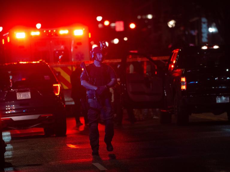 Ein mit Gewehr und Helm ausgestatteter Polizist bewacht einen Tatort im Dunkeln. Im Hintergrund sind die Lichter eines Krankenwagens zu sehen. 