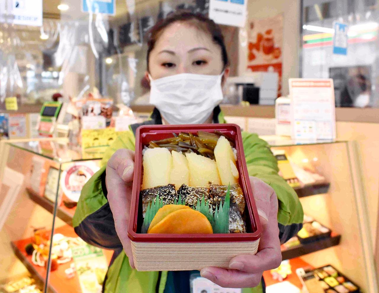 Eine Verkäuferin am Bahnhof von Hakodate in Hokkaido hält eine Box mit Essen, Bento oder auch Ekiben genannt, in die Kamera, sie trägt eine Maske.