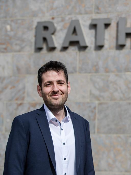 Porträt von Ryyan Alshebl (Bündnis 90/Die Grünen) - ein mittelalter Mann in Jacket und Hemd, der in die Kamera lächelt. An der Wand hinter ihm ist der Schriftzug "Rathaus" zu lesen.