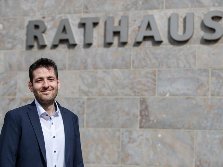 Porträt von Ryyan Alshebl (Bündnis 90/Die Grünen) - ein mittelalter Mann in Jacket und Hemd, der in die Kamera lächelt. An der Wand hinter ihm ist der Schriftzug "Rathaus" zu lesen.