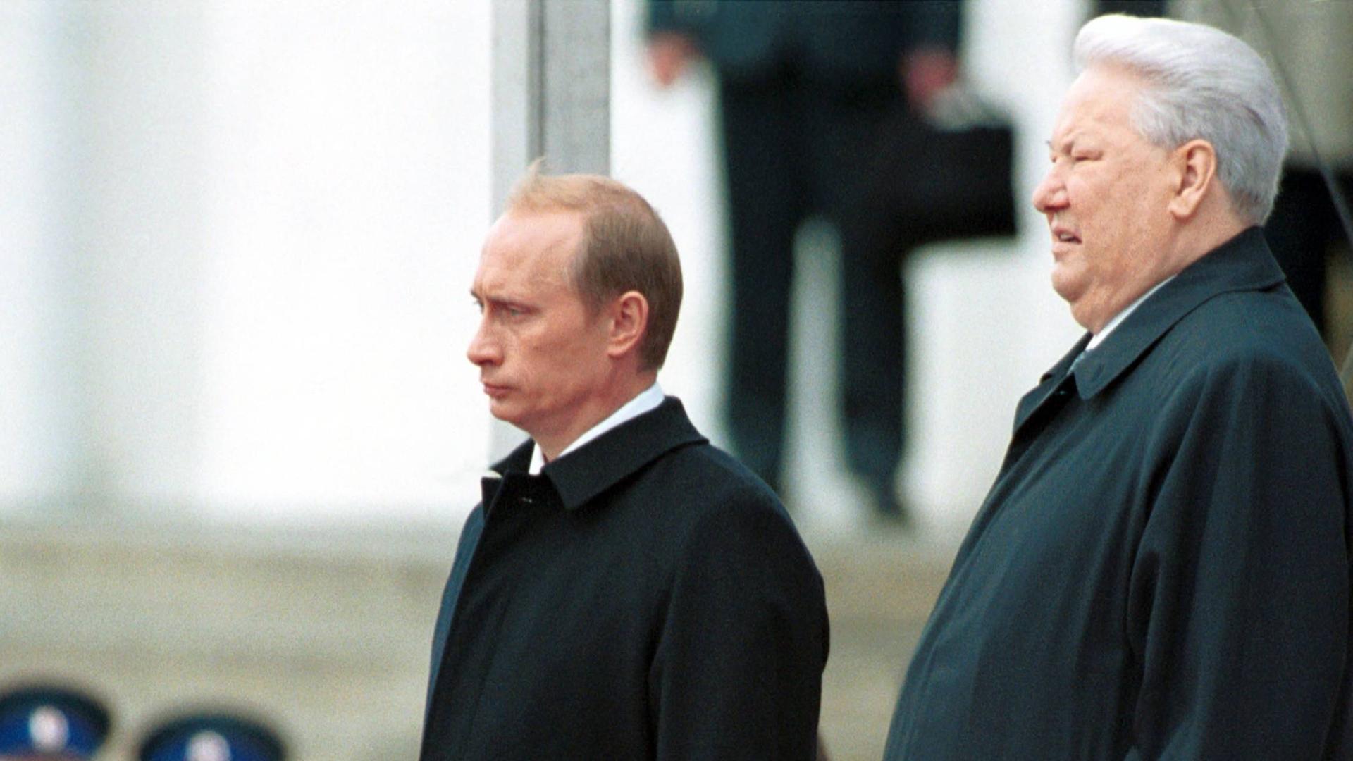 Wladimir Putin und Boris Jelzin stehen nebeneinander auf dem Kathedralenplatz im Kreml. Beide tragen dunkle Anzüge und blicken geradeaus. Die Szene zeigt die feierliche Amtsübergabe am 7. Mai 2000 – Jelzin übergibt das Präsidentenamt an Putin.