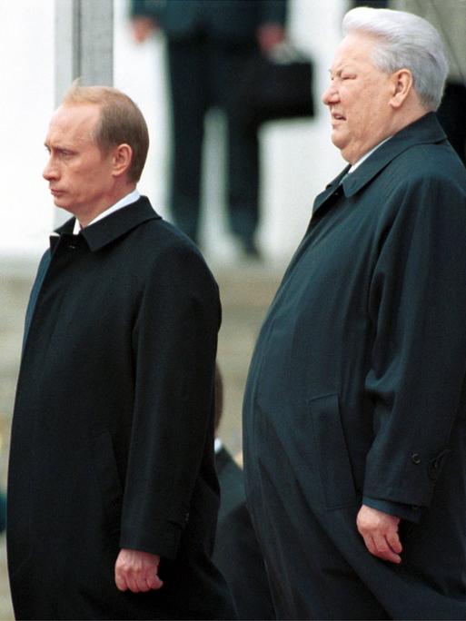 Wladimir Putin und Boris Jelzin stehen nebeneinander auf dem Kathedralenplatz im Kreml. Beide tragen dunkle Anzüge und blicken geradeaus. Die Szene zeigt die feierliche Amtsübergabe am 7. Mai 2000 – Jelzin übergibt das Präsidentenamt an Putin.